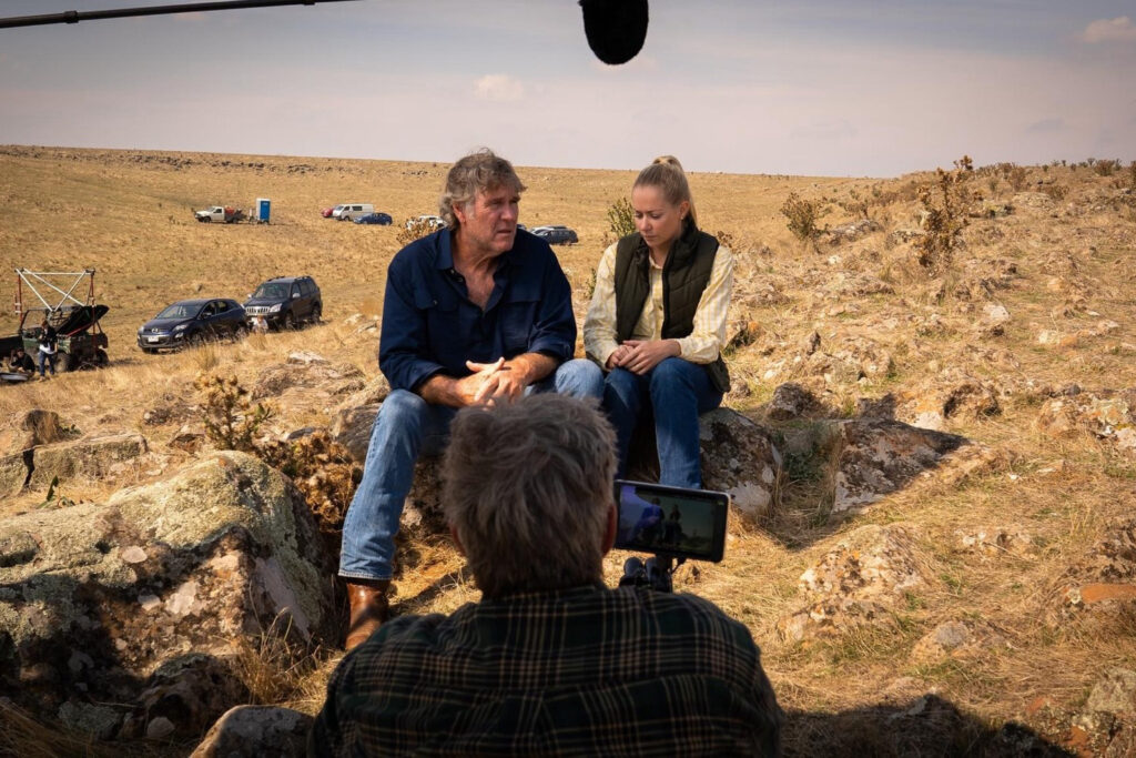 A man and a woman acting in a scene being filmed on a hill in a rural setting.