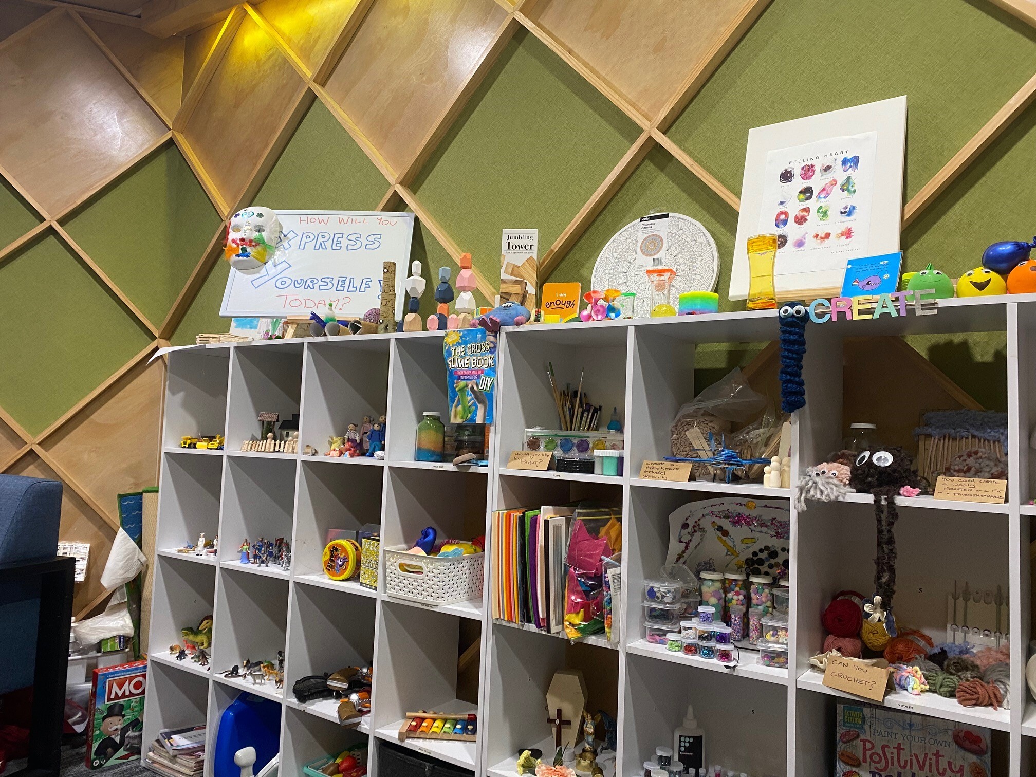 A bookshelf full of toys and games in front of a green decorated wall.