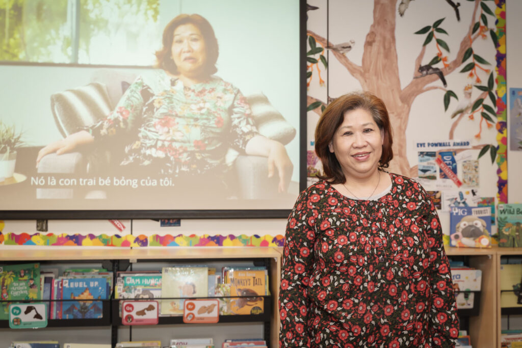 A carer named Theresa standing in front of a projected image of herself on film in a room with a painted wall and childrens books on a shelf.