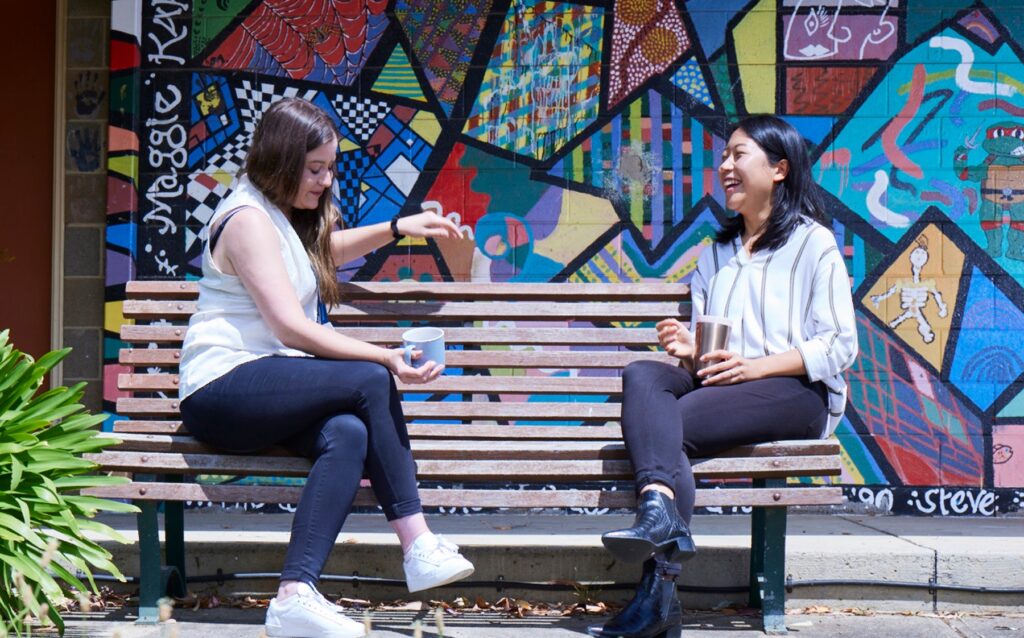 Two women sitting on a park bench drinking coffee in front of a colourful wall