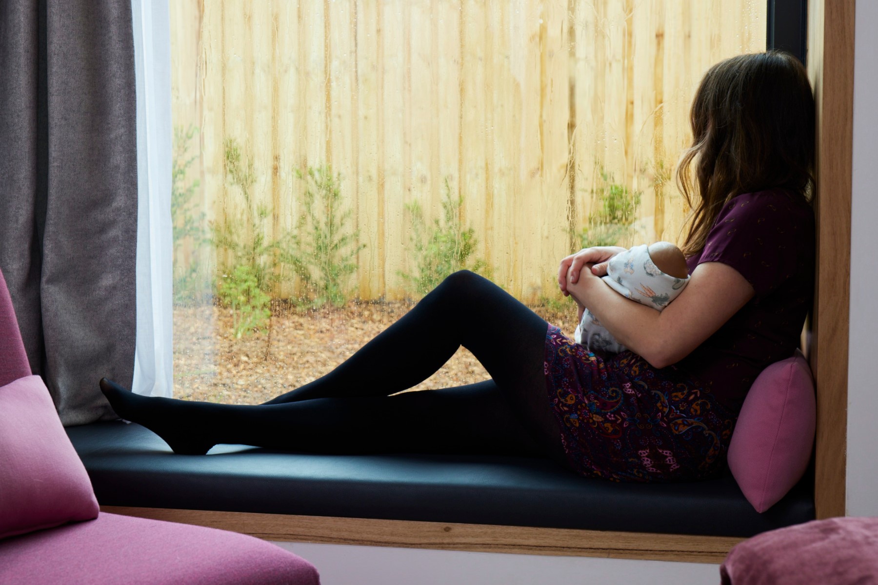 A woman holding a baby sitting in a window frame looking out at a garden