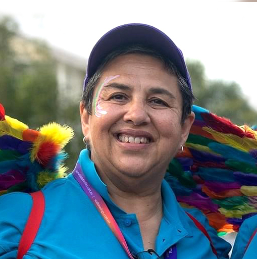 A portrait of a lady smiling at a pride parade wearing colourful face paint and rainbow feather wings
