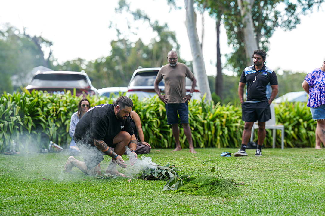A person kneeling on green grass handling a smouldering branch of gum leaves