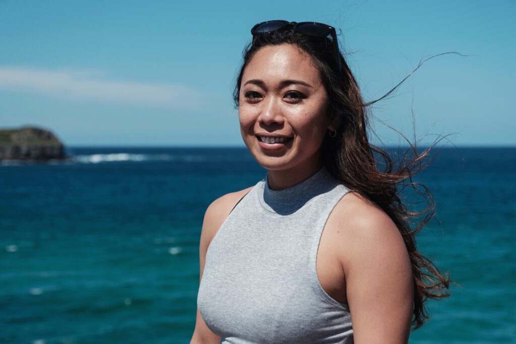 A young Asian woman smiling toward the camera with the ocean in the background.