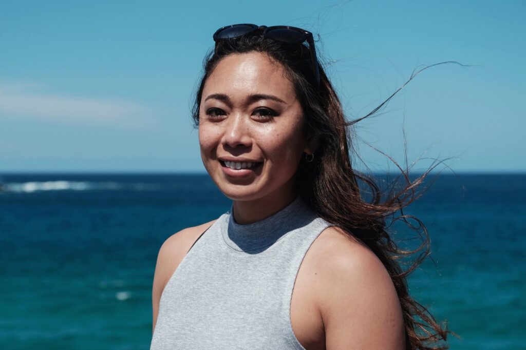 A young person with long dark hair smiling toward the camera with the ocean in the background