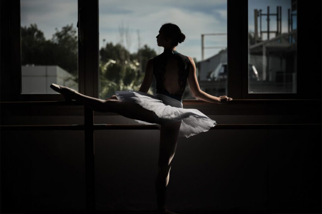 Back view of slender ballerina standing at ballet barre and stretching raised leg during rehearsal in evening