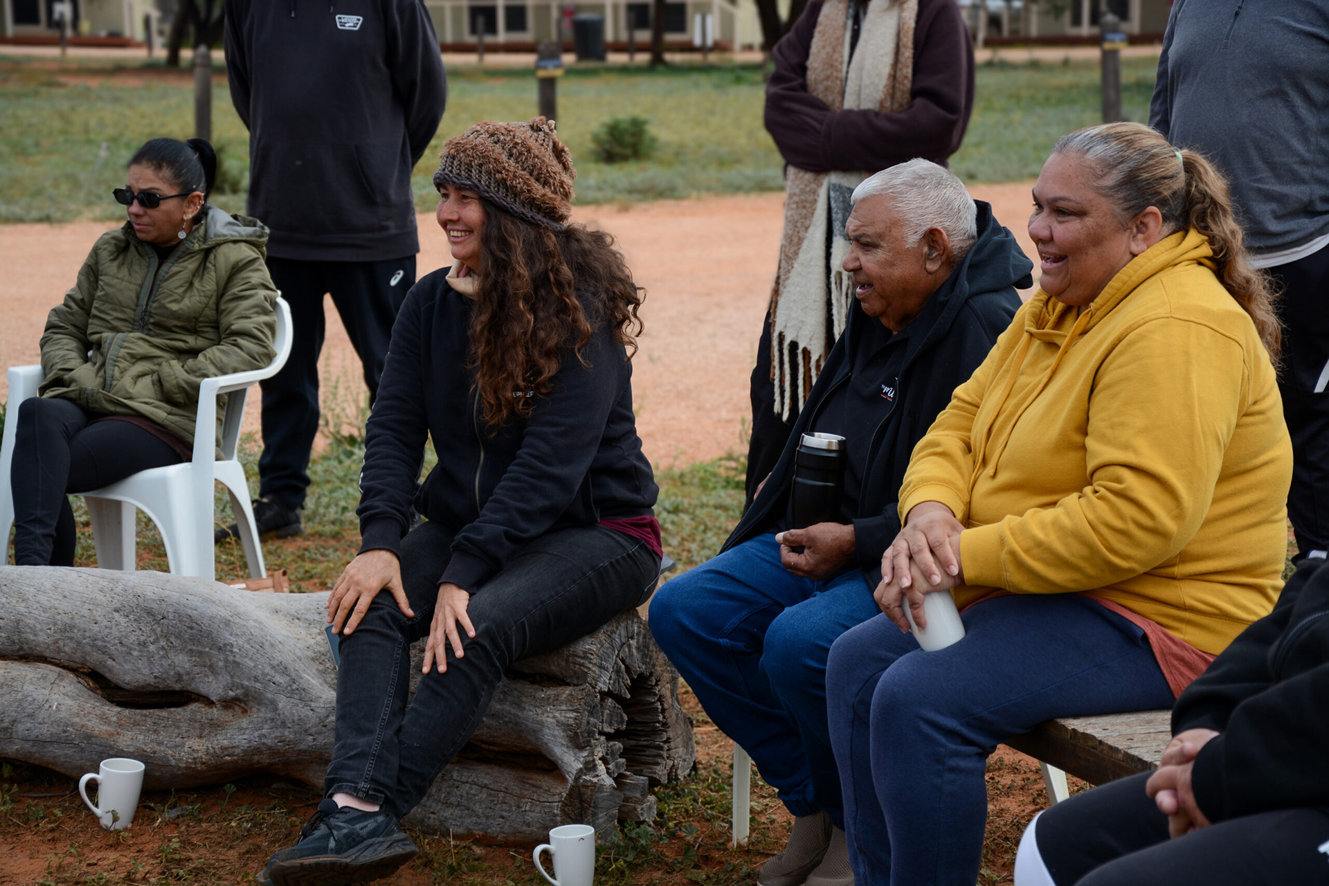 People wearing warm clothing, sitting on wooden benches and an old tree log laughing and chatting outside