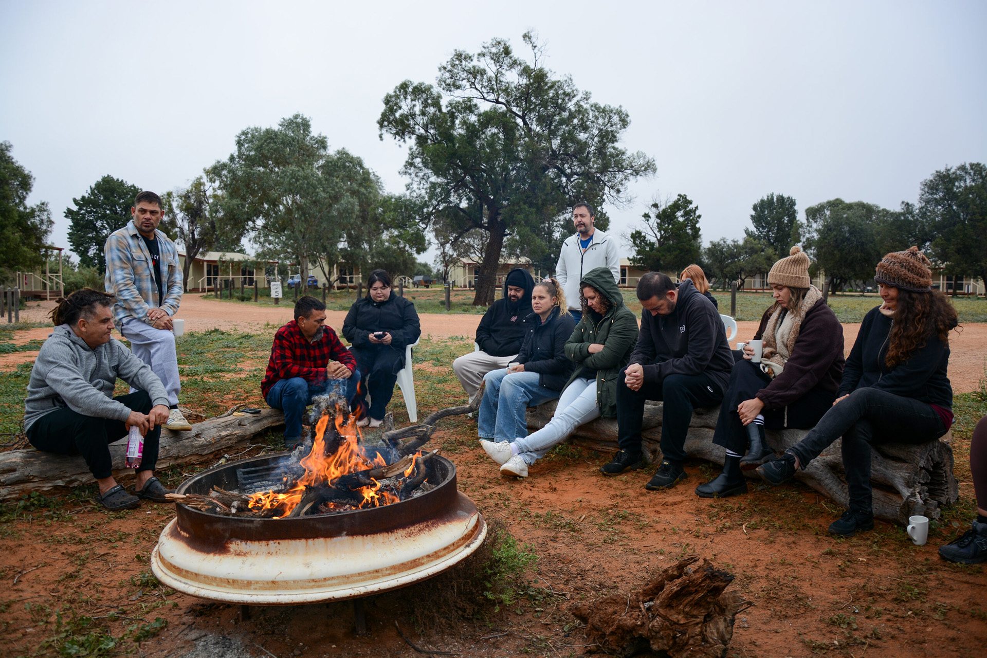 A group of people wearing warm clothing, sitting around a large fire pit drinking tea and chatting