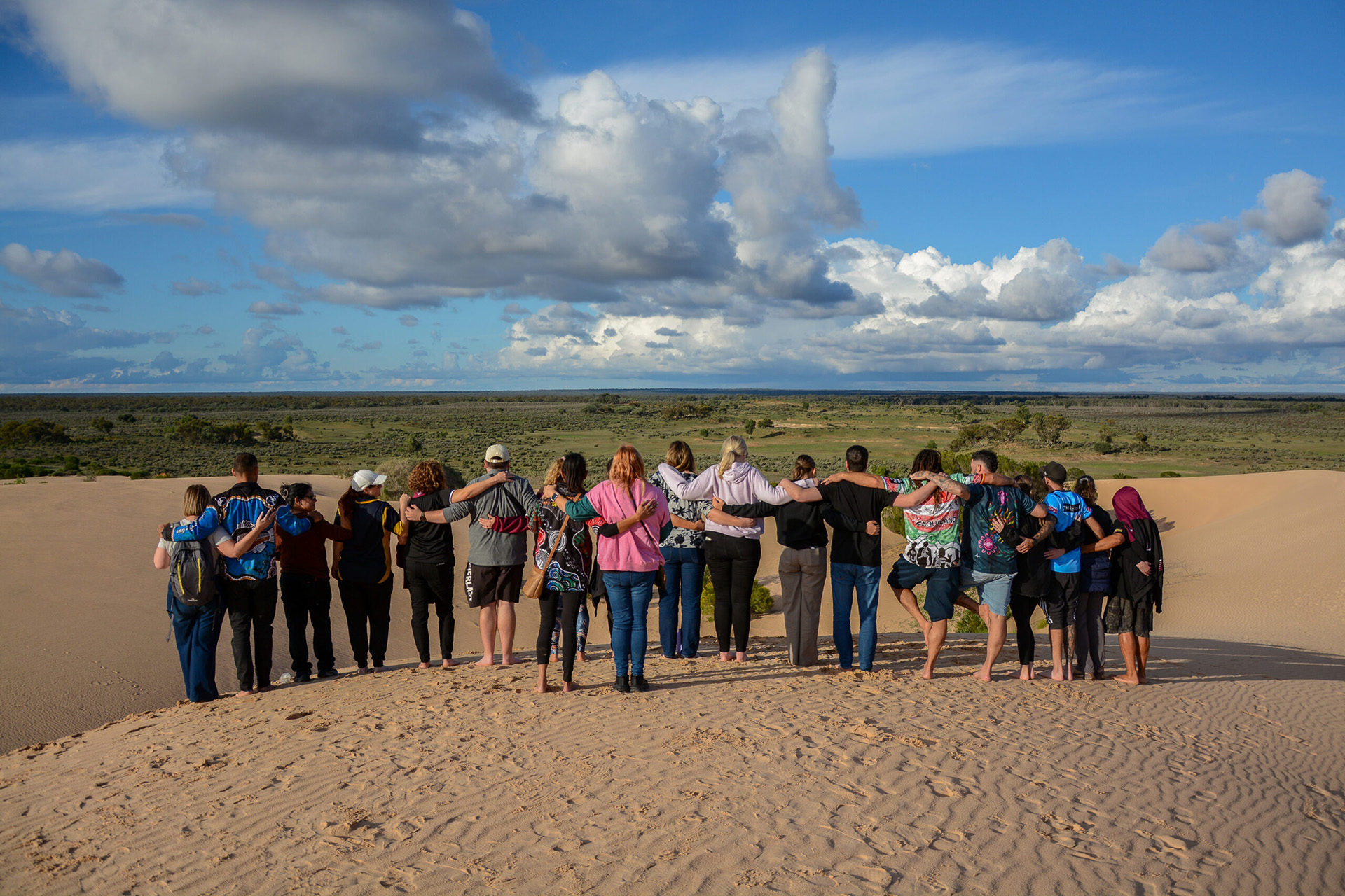 A large group of people, arm in arm, standing at the top of a sand dune, looking out across a vast desert landscape