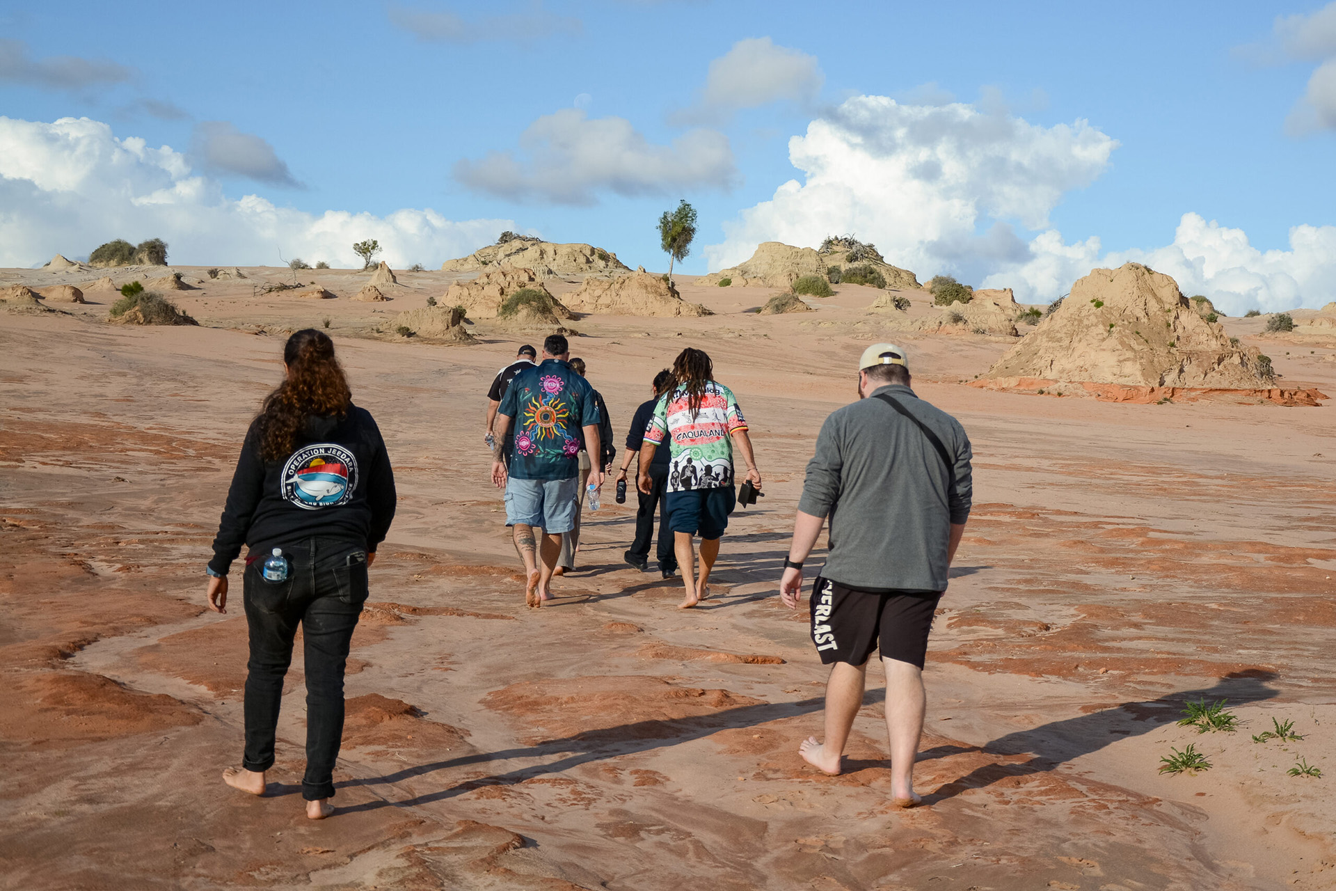 Seven people walk barefoot on red dirt, towards a sandy hill with large protuding rock formations