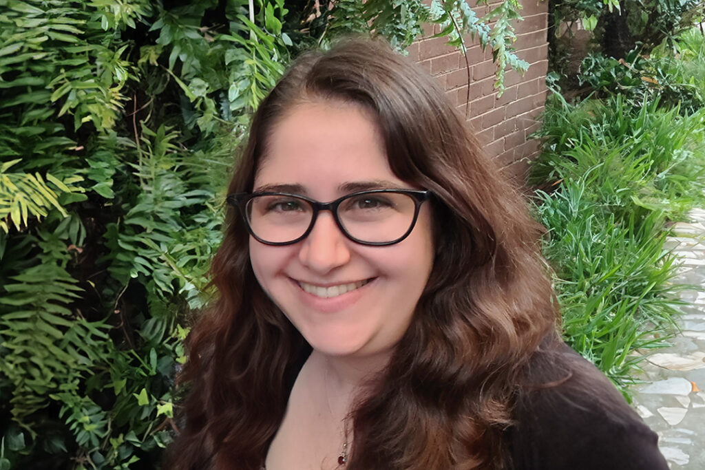 A person with long brown hair, wearing glasses, smiling in front of a wall covered in green ferns.