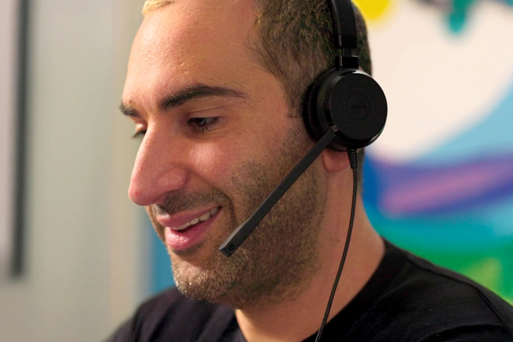 A close up of a person with short bleached blonde hair wearing a headset in front of a colourful wall