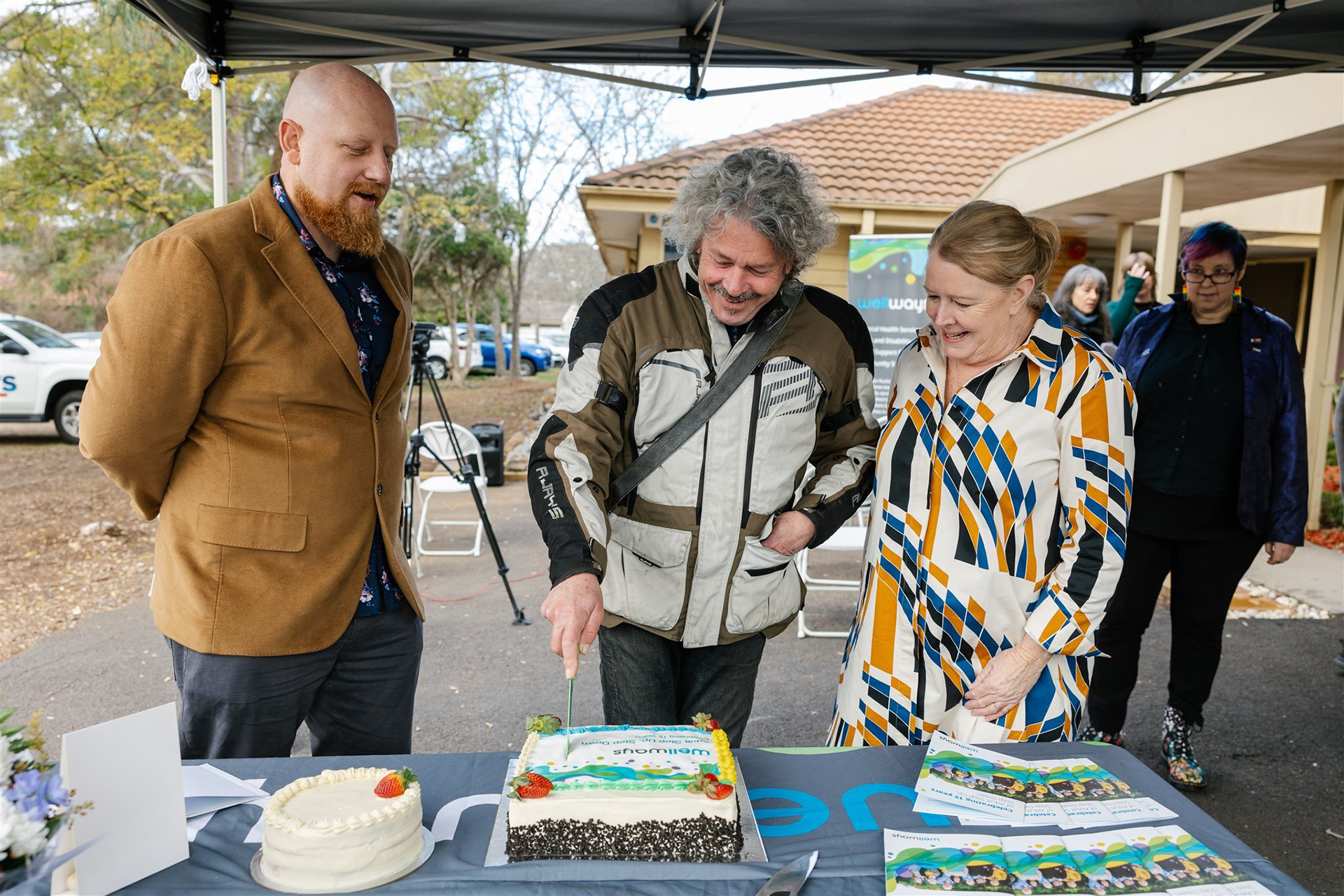 Three people standing under a marquee in a carpark cutting a cake during an outdoor event.