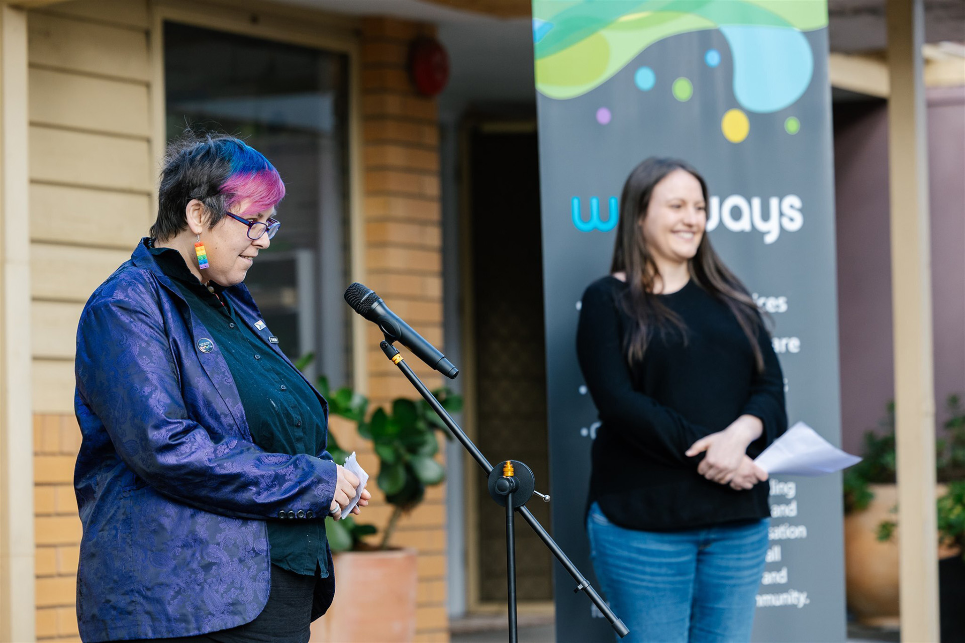 A person with colourful hair and a purple jacket is speaking to a microphone while another person stands behind her in front of a Wellways pull-up banner.