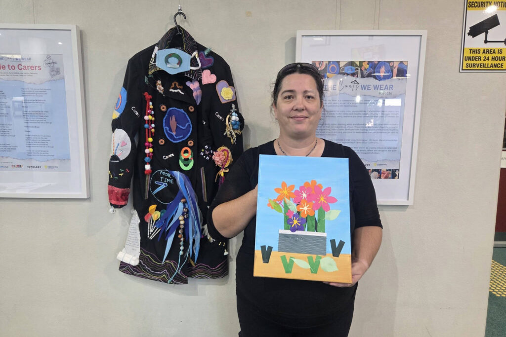 A person with dark hair and dark clothing holding a painting of a flower bouquet, standing next to a coat covered in trinkets hanging on a wall