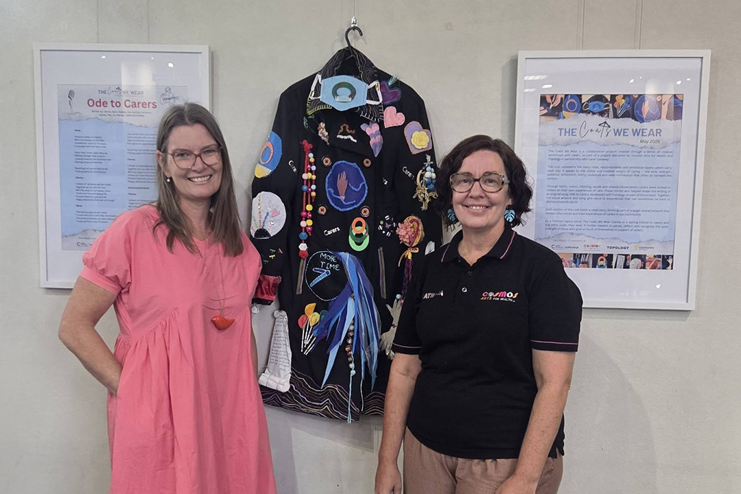 Two people standing in front of a wall where a dark coat covered in decorations is hanging.