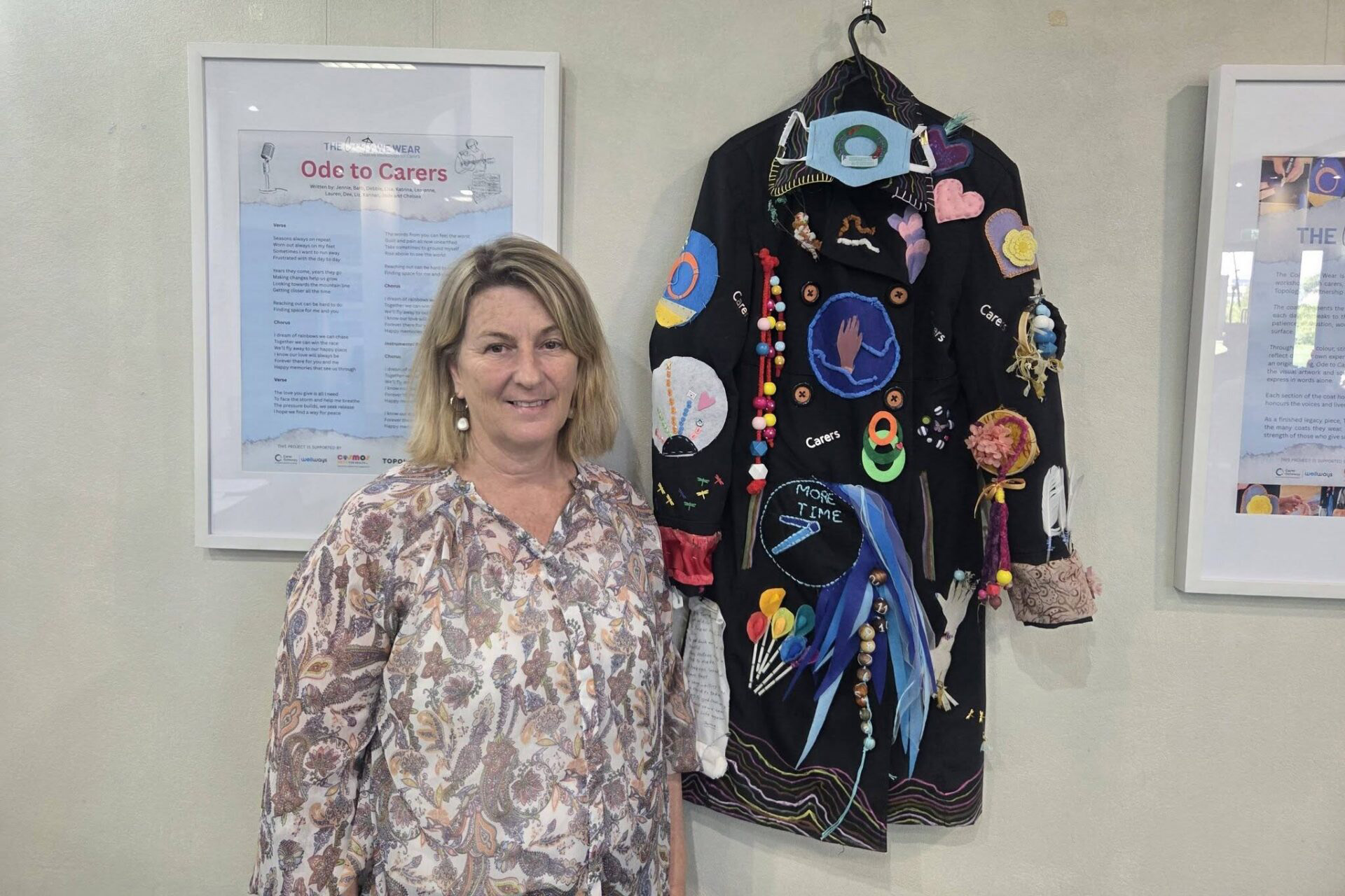 A person with medium length hair and a floral shirt standing next to a coat covered in trinkets hanging on a wall.