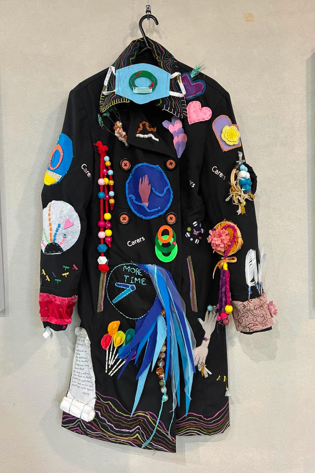 A dark coat, covered in colourful trinkets hanging on a wall.