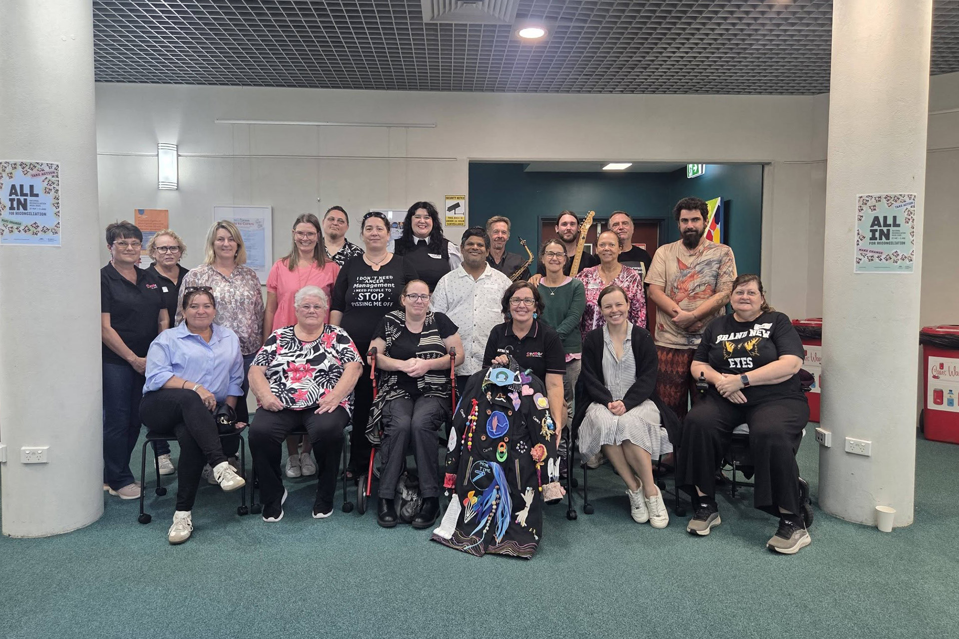 A large group of people in a room or hall. Some people are standing at the back and a row of people in seats and wheelchairs at the front.