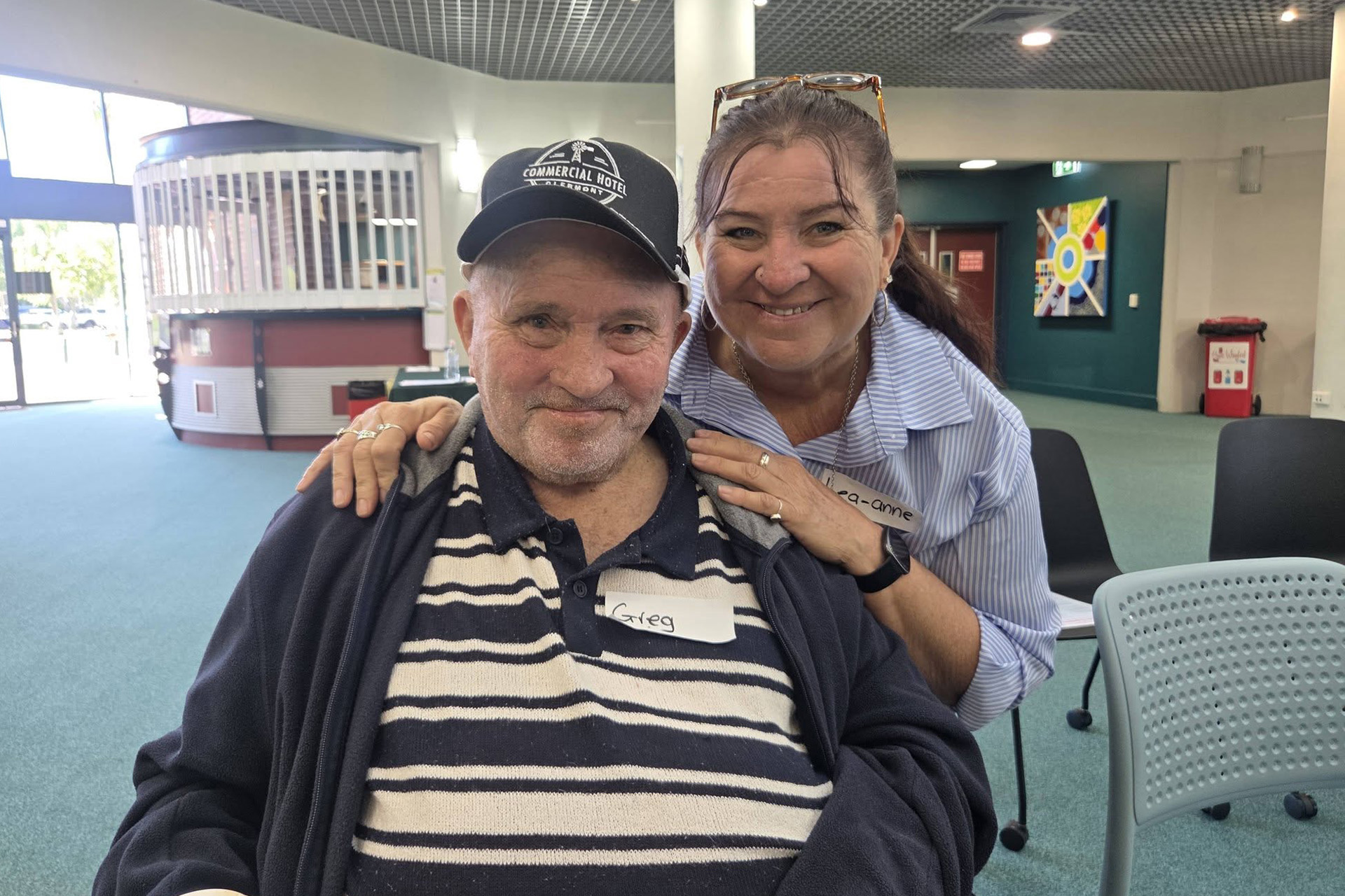 An elderly person in a hat and a striped shirt, smiling and sitting in a wheelchair. Their carer is crouching behind them with their hands on their shoulders, smiling.
