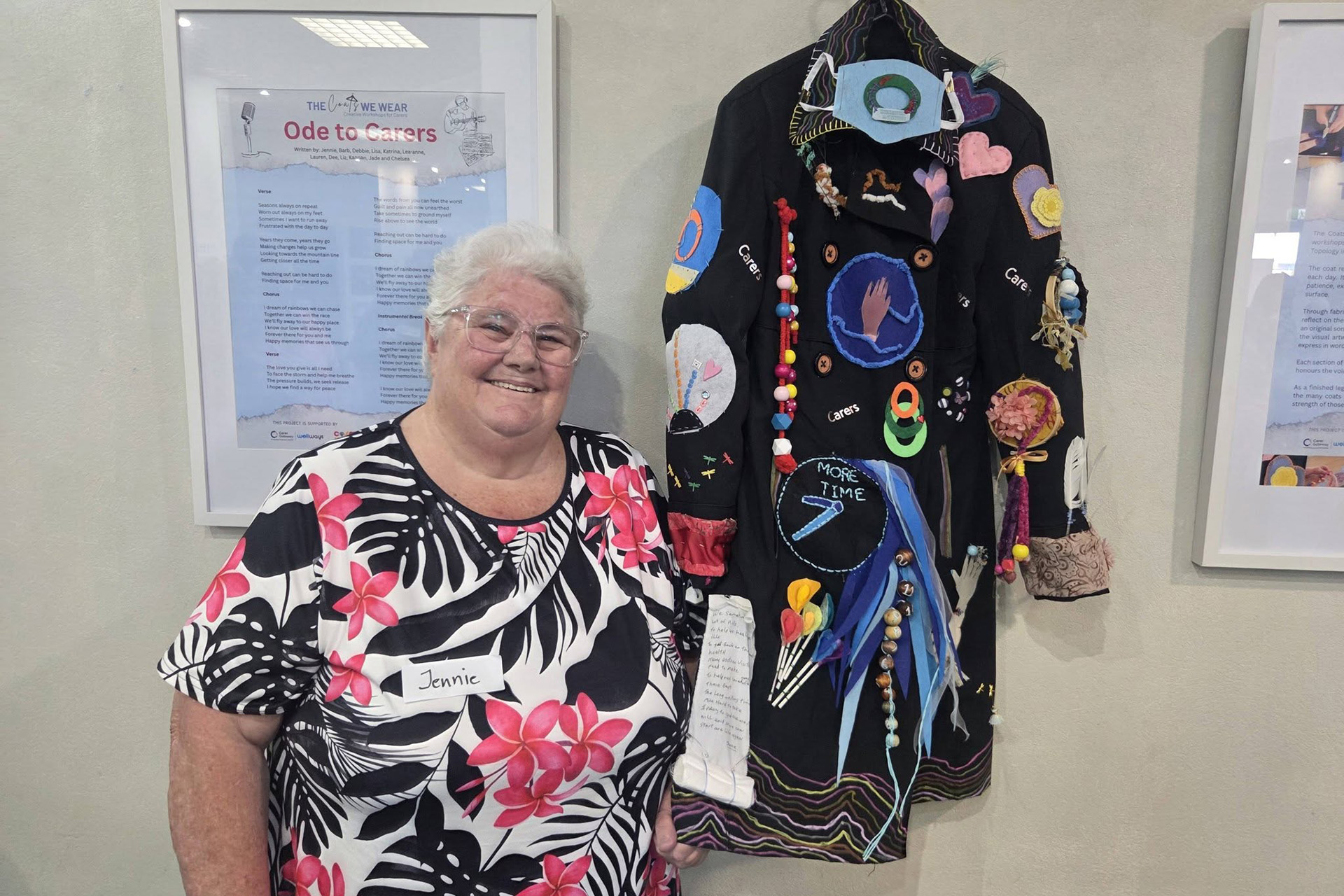 A person with short white hair, leaning against a wall where a dark coat covered in decorations is hanging.