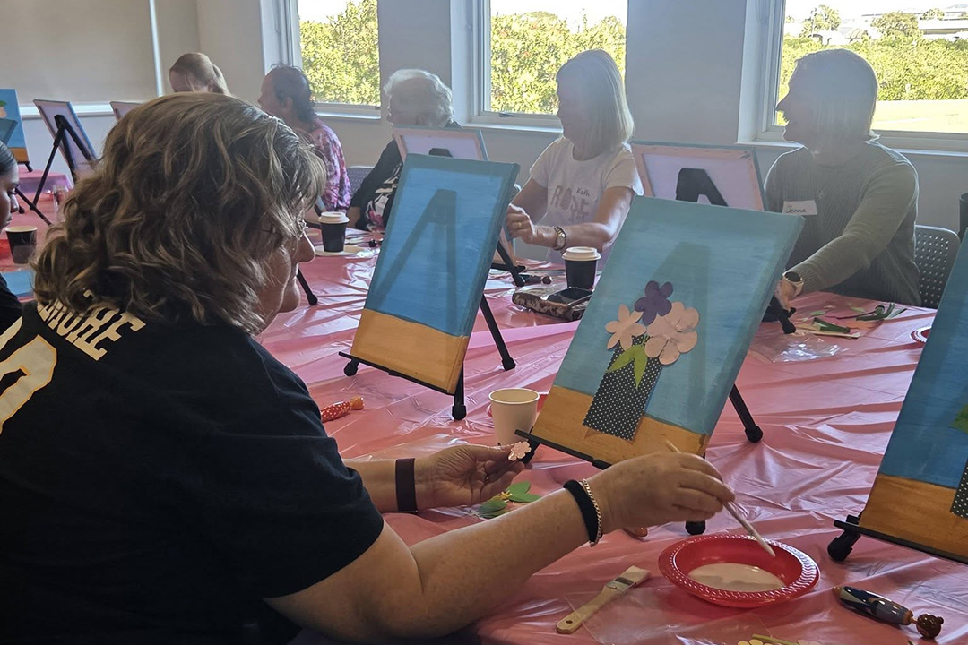 A large group of people seated at a long table painting on canvas held by small Eisel stands.