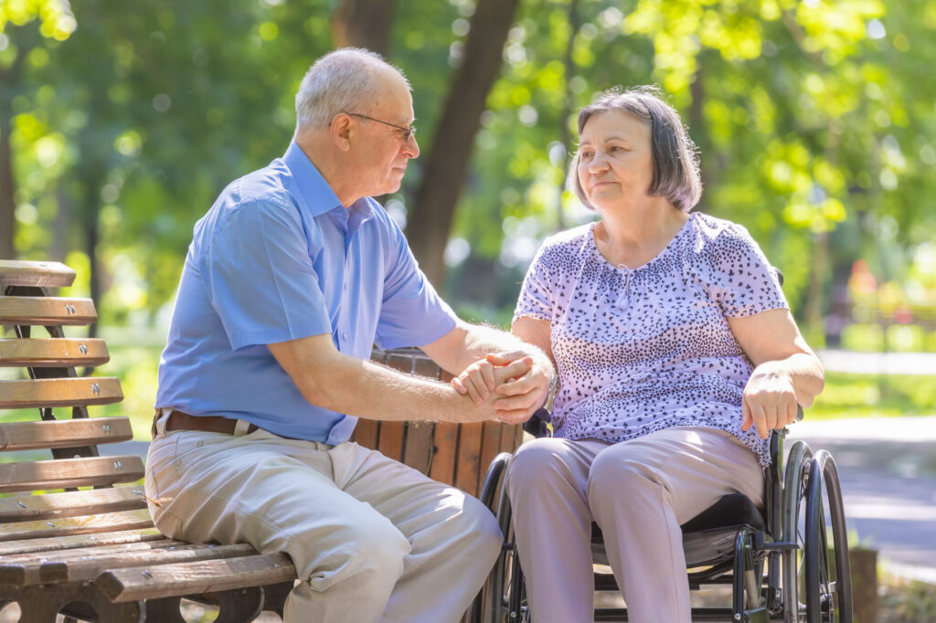 A person sitting on a park bench gently holding the hand and looking into the eyes of their loved one in a wheelchair