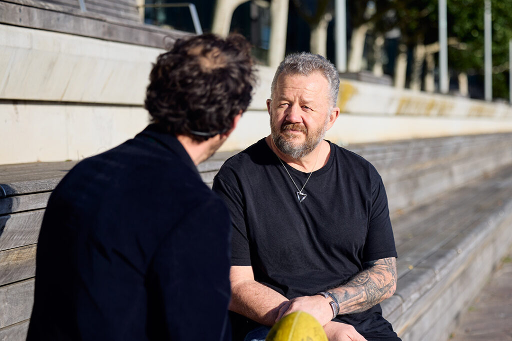 Two people wearing black clothing sitting on outdoor steps talking. One is holding a yellow football.