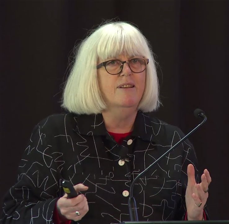 Mary O'hagan speaking at a conference in front of a black background.