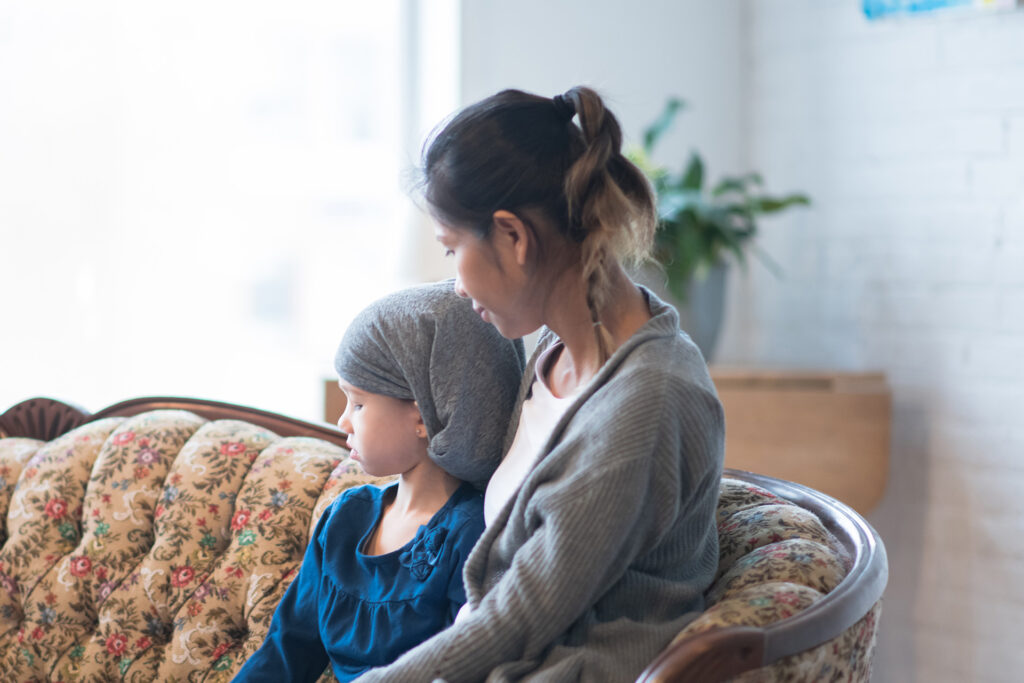 A person with long dark hair and a grey top, sitting on a couch with their arm around a child wearing a bandana after having chemotherapy.