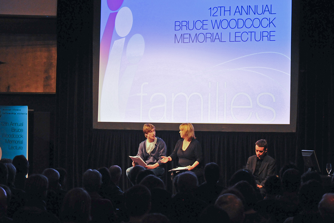 Audience watching a panel discussion at the 12th Annual Bruce Woodcock Memorial Lecture, with two speakers seated on stage in front of a large screen displaying the event title and the word 'families' in the background.