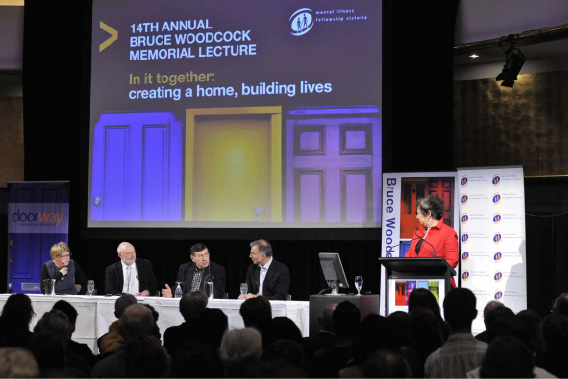 Audience watching a panel discussion at the 14th Annual Bruce Woodcock Memorial Lecture, with four speakers seated on stage in front of a large screen displaying the event title and one speaker behind a lectern.