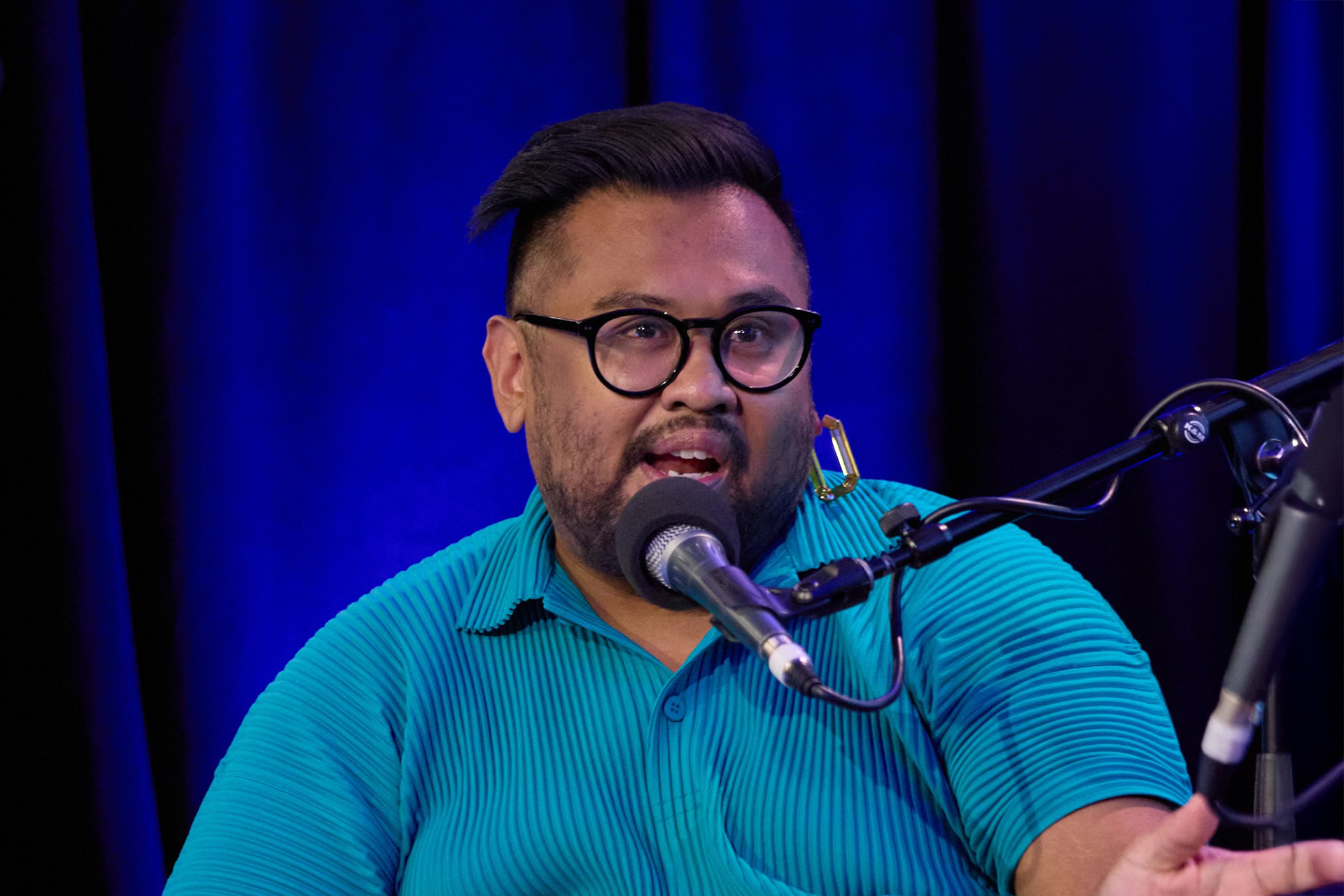 A person in a green shirt and black framed glasses, speaking to a microphone in front of a blue stage curtain. 