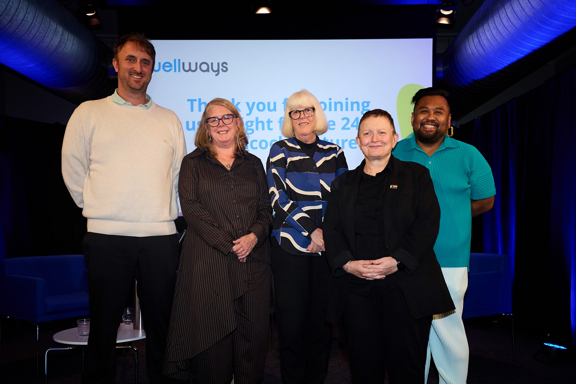 Five smiling people stand on stage side by side in front of a projector screen. 