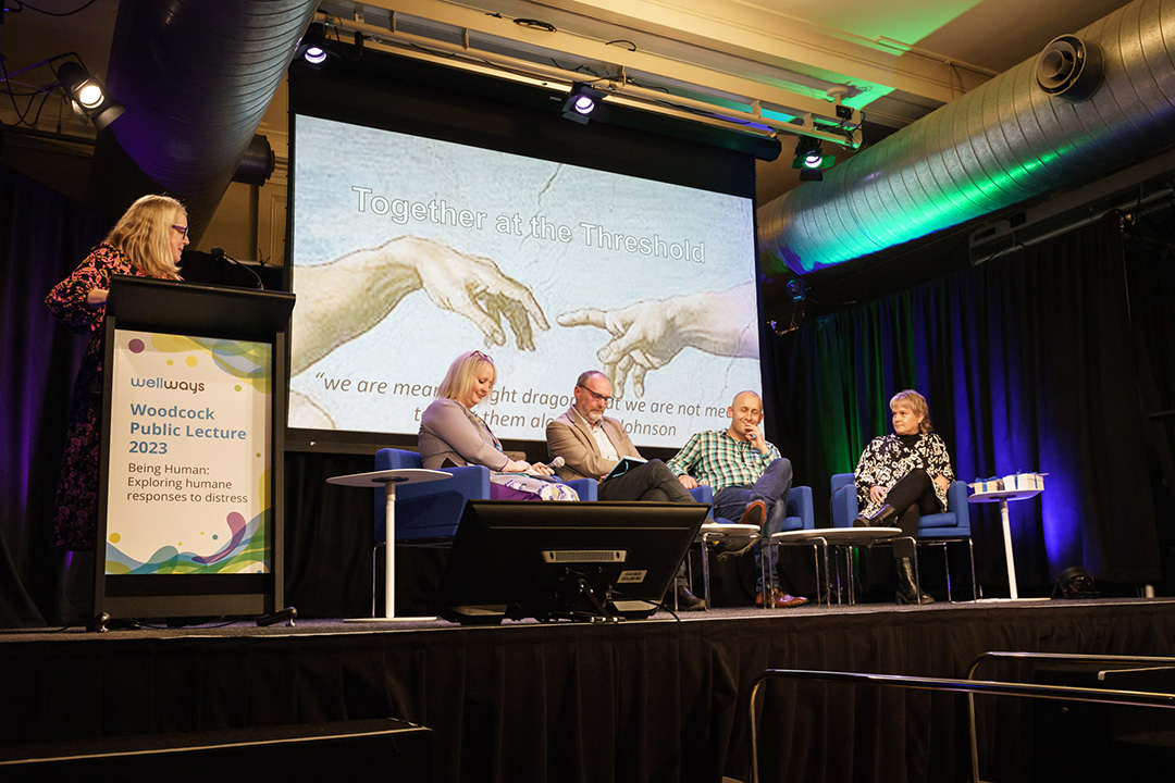 Four speakers seated on stage in front of a large screen displaying hands and the words 'Together at the Threshold' in the background. One person stands off to the left side behind a lectern with the event title 