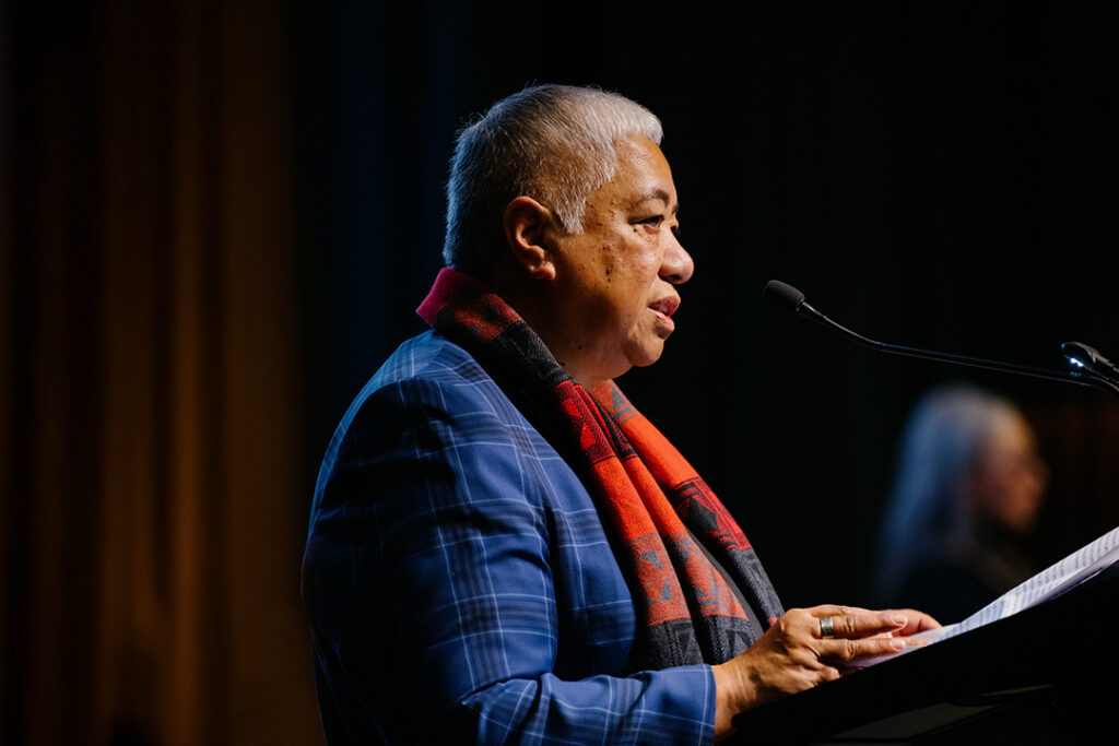 A speaker in a blue plaid coat and red plaid scarf stands at a lectern under a spotlight, speaking into a microphone.