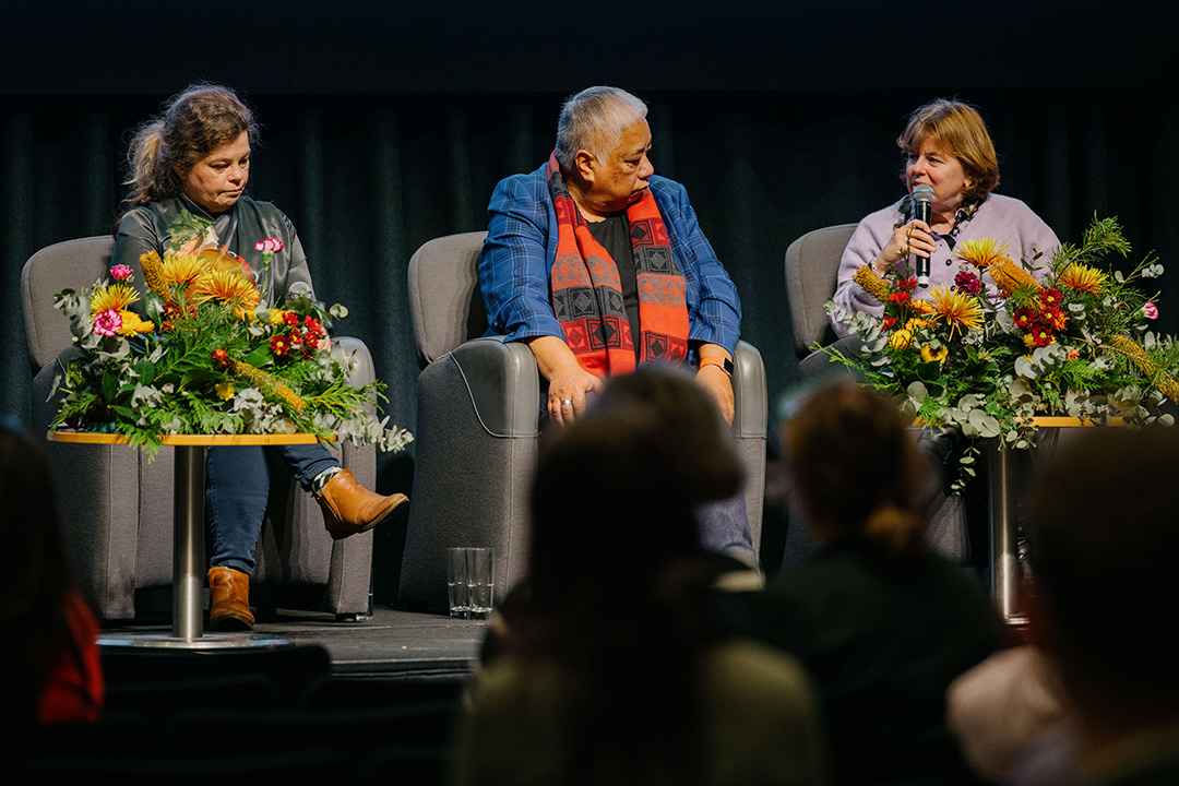 Three people sitting on a stage in front of a black curtain speaking. There are two small tables with large colourful flower arrangements.