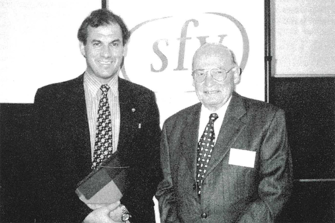 An old looking grayscale photograph of two people wearing suits standing in front of a sign with the letters 