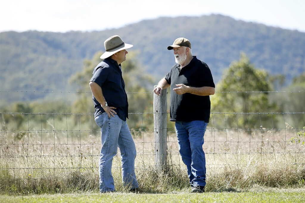 Two people wearing hats standing next to a wire fence talking in-front of a bushy hill.
