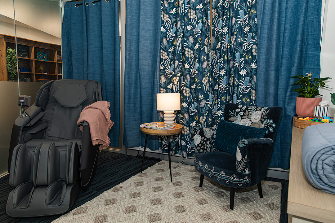 A room with long blue curtains, cream carpet, a massage chair and a blue velvet armchair.