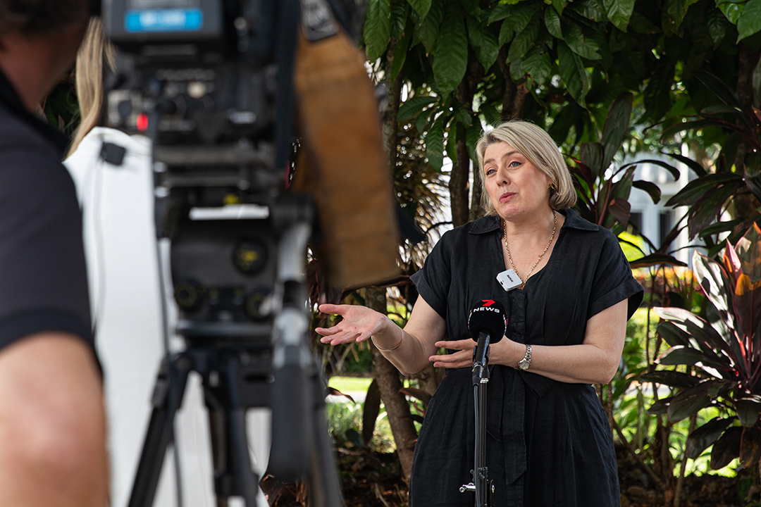 A person standing in front of a garden speaking to a microphone in front of a camera.