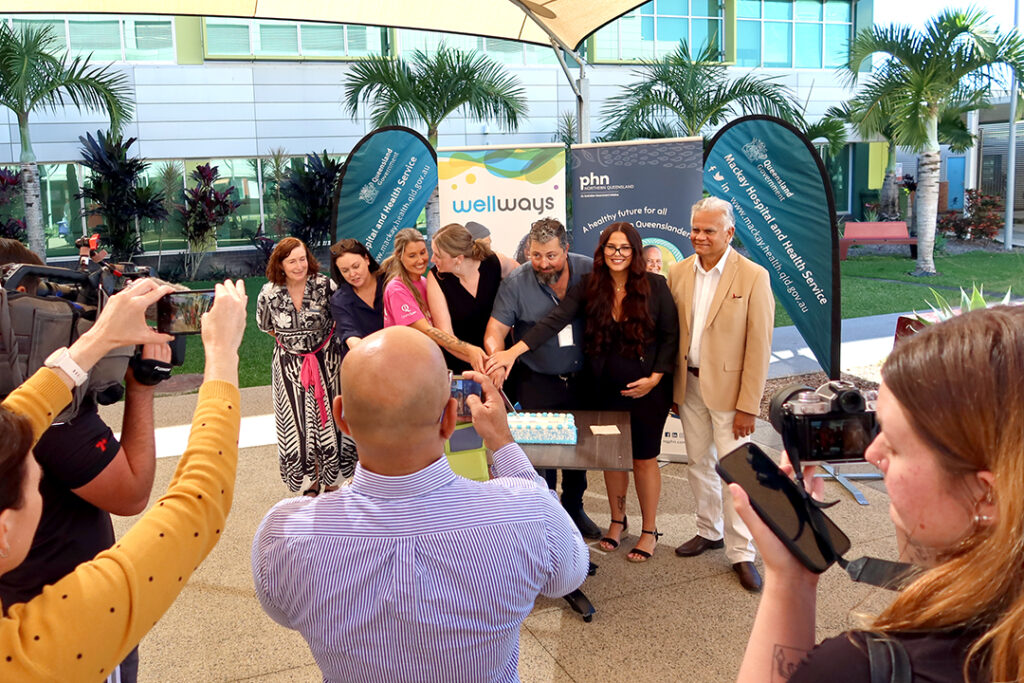 A group of people in front of signage cutting a cake with people taking photos in the foreground.