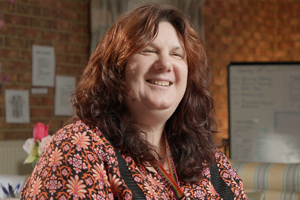 A person with long dark red hair and a floral print shirt in a room with brick walls.