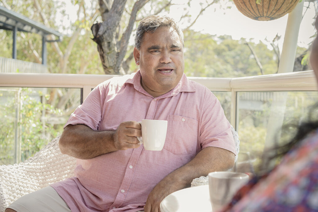 A person wearing a light red shirt drinking a coffee on a balcony surrounded by trees.