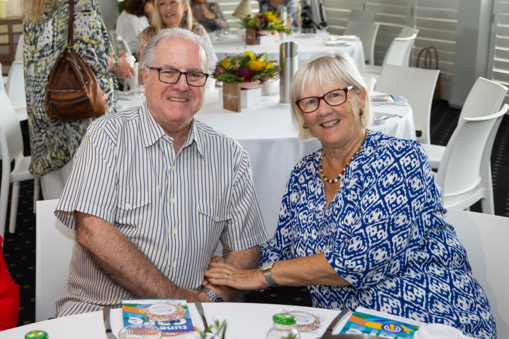 Two older people sitting down at a table at an event holding hands and looking happy