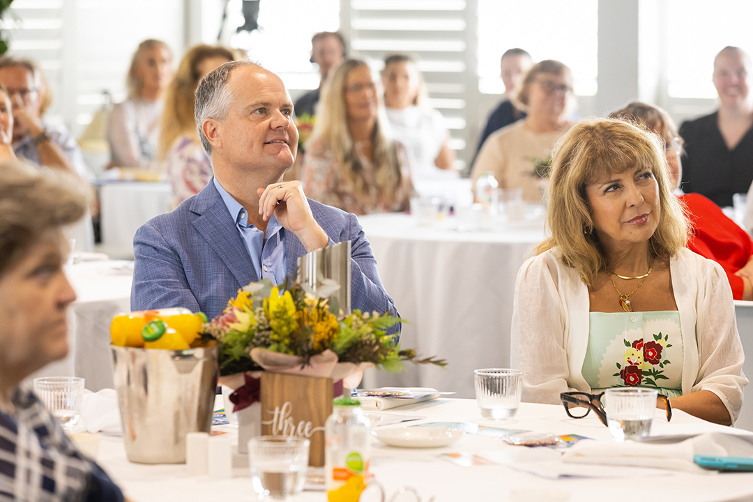 A room full of people sitting at tables with white table cloths and floral arrangements