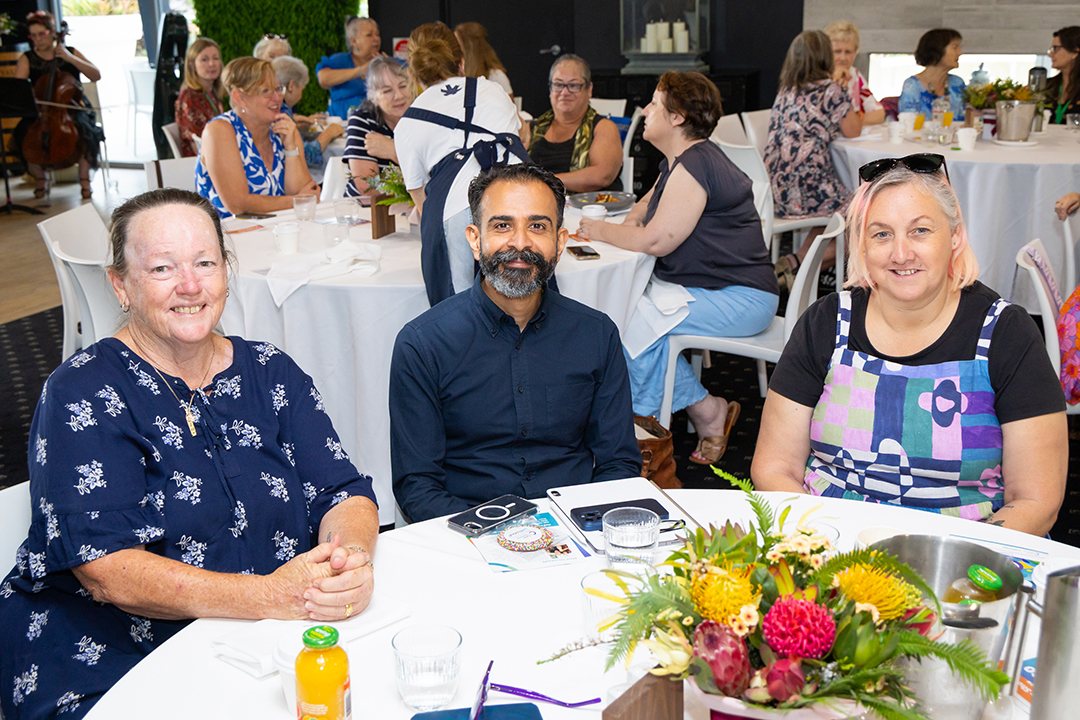 Three people sitting and smiling at a table with a bright coloured floral arrangement