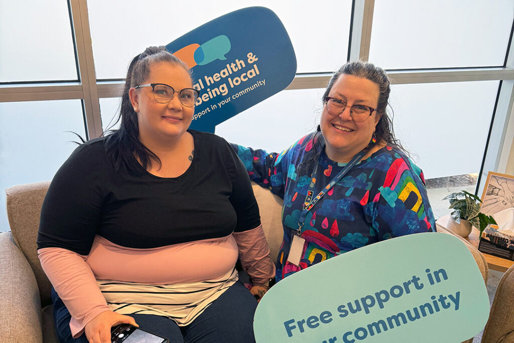 Two people sit on a sofa. One is holding large blue signs in each hand.