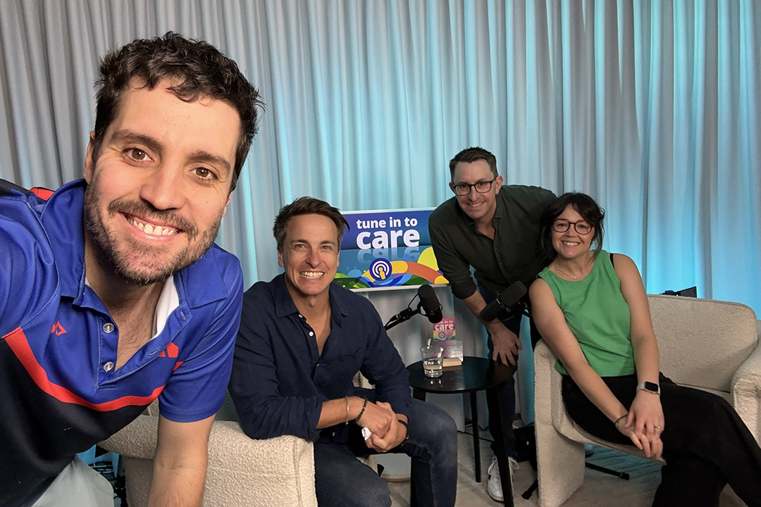 A group of people. Some standing, some sitting on white lounge chairs in a room with a curtain, microphone and blue lights
