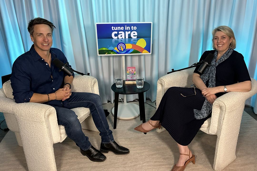Two people sitting on white lounge chairs in a room with a curtain, microphone and blue lights
